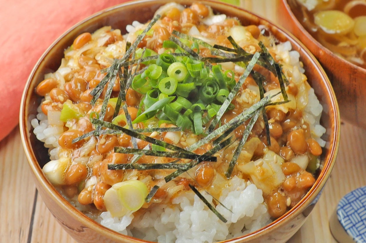 ご飯の上に納豆、刻み海苔、刻みネギがのった丼と味噌汁のセットが木のテーブルに置かれている。