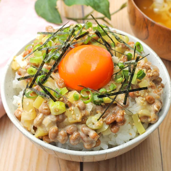 ご飯の上に納豆、卵黄、海苔、小ねぎが乗った和風丼。背景には味噌汁も見える。