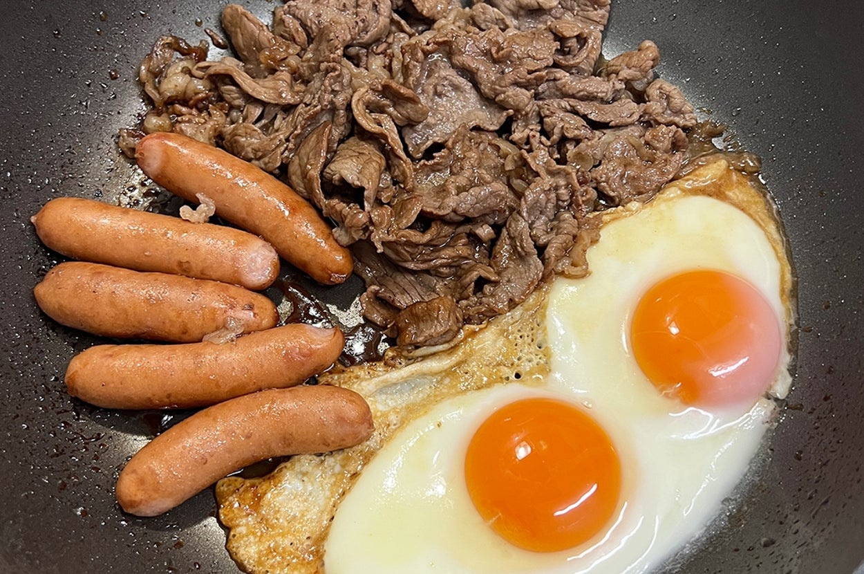 ソーセージ、薄切り牛肉、目玉焼き2つがフライパンで調理されている様子。