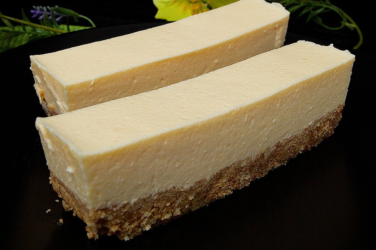 2本のチーズケーキが黒い皿に乗っている。背景には黄色い花と緑の葉が飾られている。