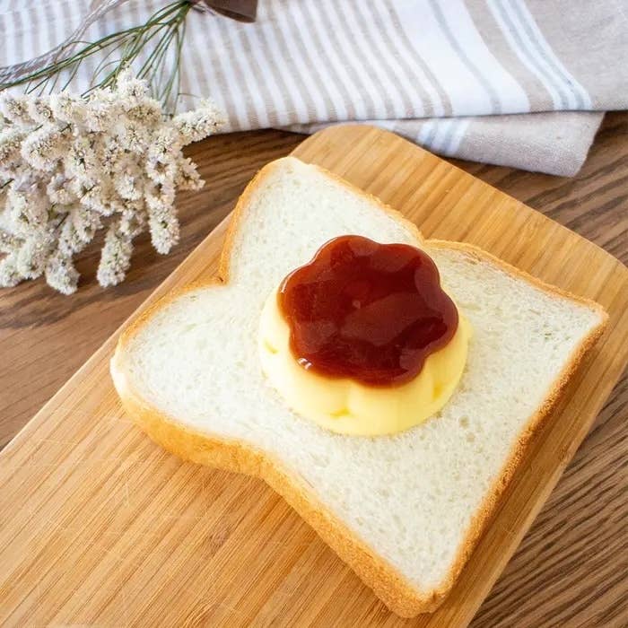 食パンにプリンといちごソースがのったユニークなデザート。木製のプレートの上に置かれ、背景にタオルと花がある。