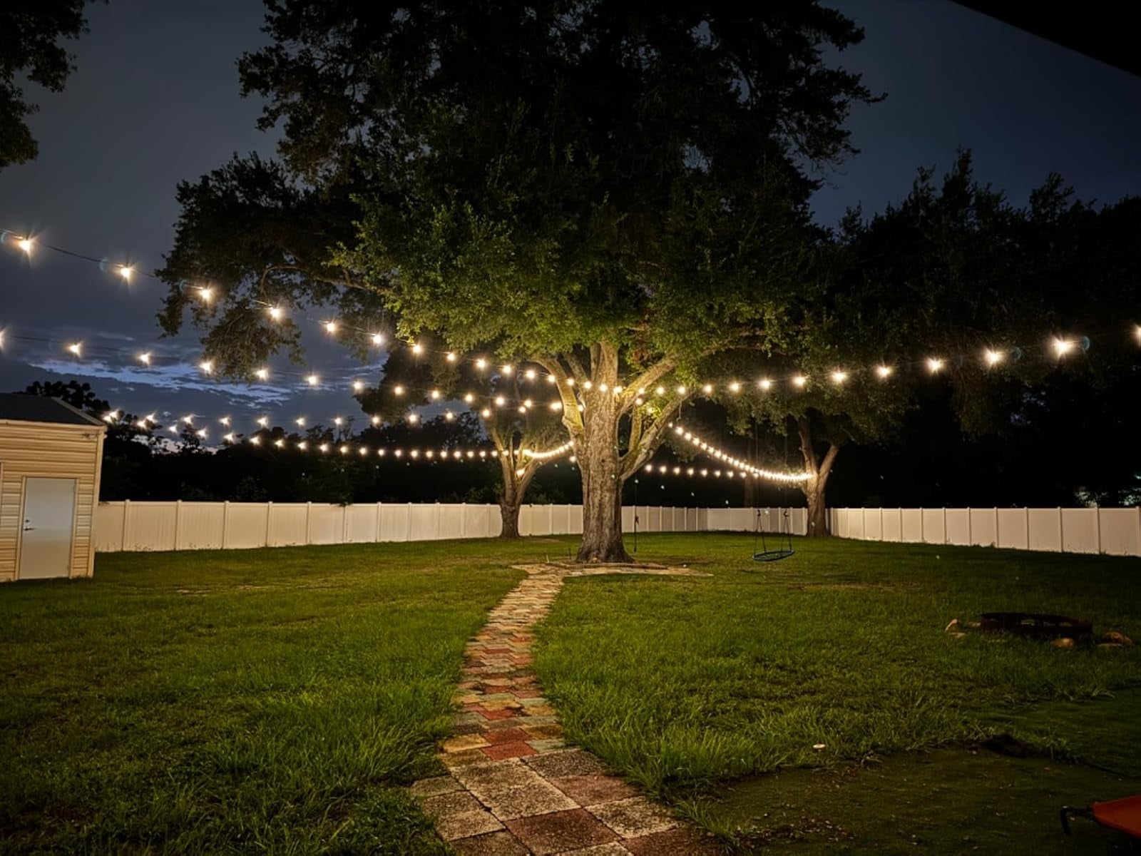 A backyard with drawstring  lights hanging betwixt  trees, illuminating a walkway. Enclosed by a gangly  fence, cleanable   for outdoor gatherings