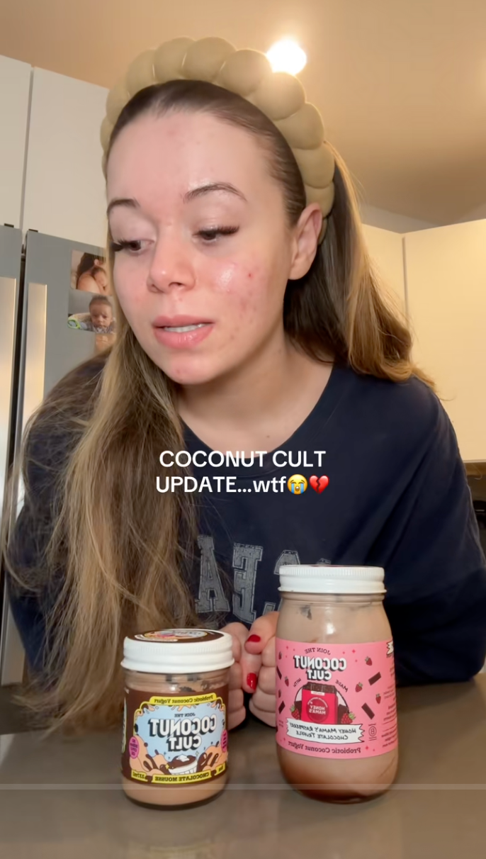 A person with a headband looks at two jars of Coconut Cult products on a kitchen counter. Text overlay reads: "COCONUT CULT UPDATE...wtf."