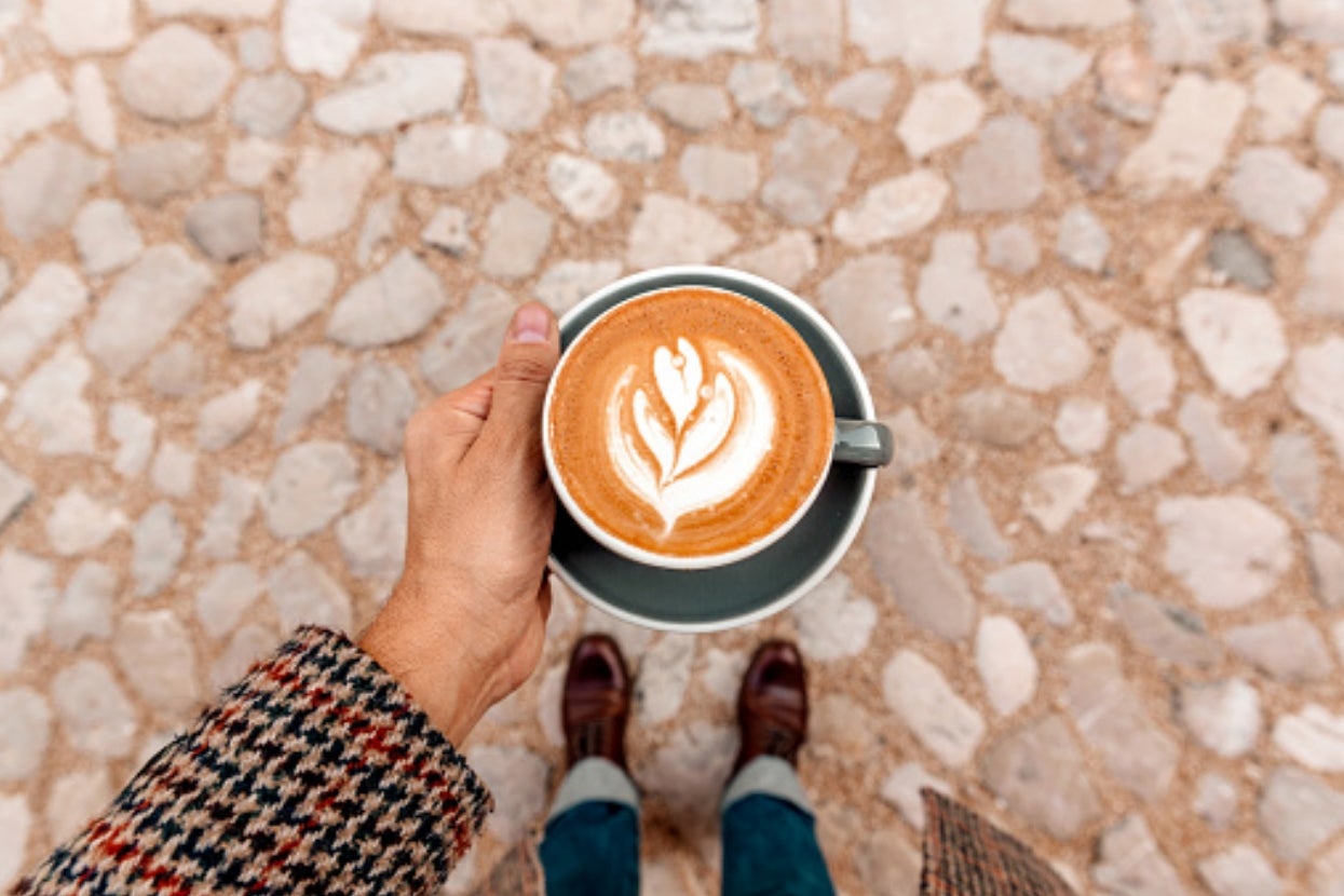 Person holds a latte with heart-shaped foam art while standing on a cobblestone path, wearing a houndstooth coat and brown shoes