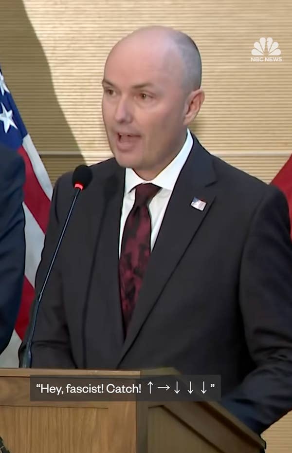 A person in a suit speaks at a podium with flags in the background. Subtitles show: “Hey, fascist! Catch! ↑→↓↓↓”
