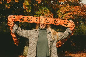 Person holding a string of paper jack-o'-lanterns in front of their face, smiling with autumn leaves in the background