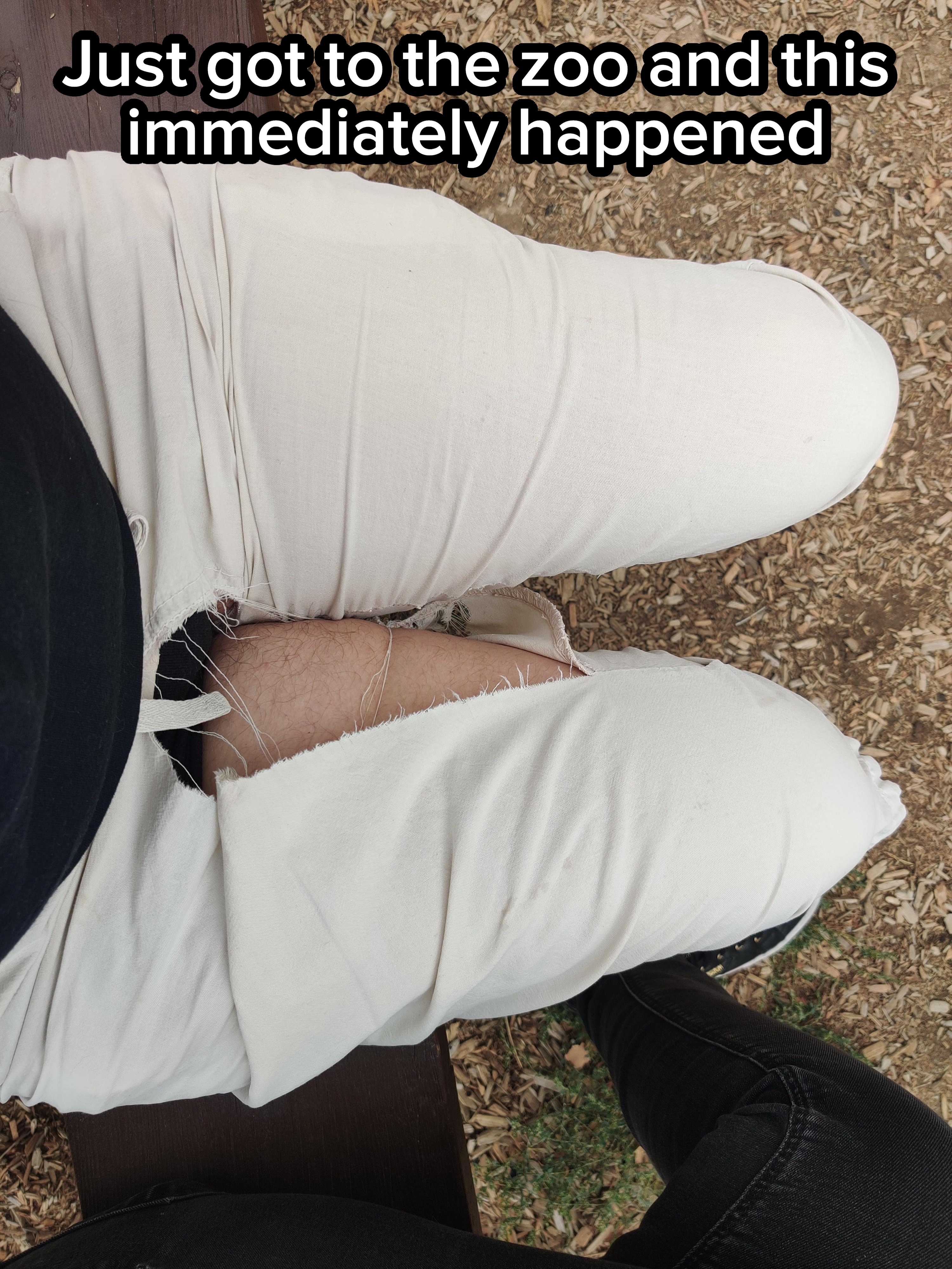 Pants ripped at the seam near the knee while sitting on a bench, exposing skin underneath. Person wears dark top and dark footwear