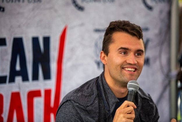 A person smiles while holding a microphone at an event with a large partially visible banner behind them