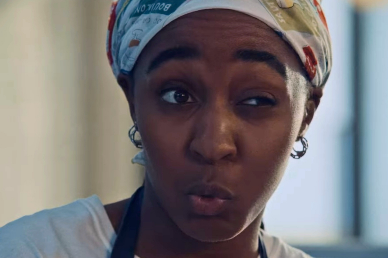 Person wearing a headscarf and apron makes a playful facial expression in a kitchen setting
