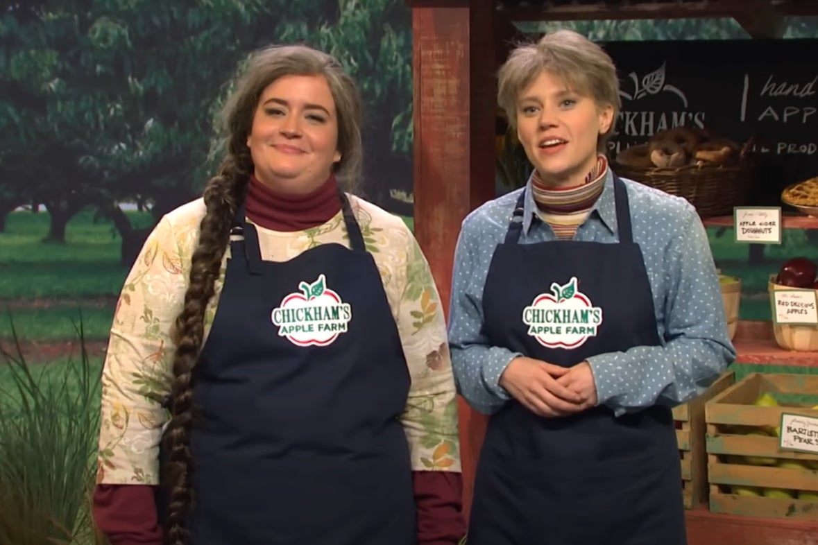 Aidy Bryant and Kate McKinnon in "SNL" skit wearing "Chickham's Apple Farm" aprons standing in front of a farm produce display, smiling