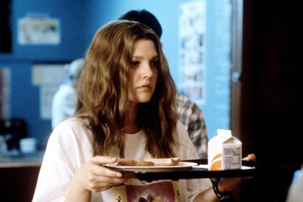 Drew Barrymore in "Never Been Kissed" with long wavy hair holds a cafeteria tray with food and beverage, appearing focused in a casual indoor setting