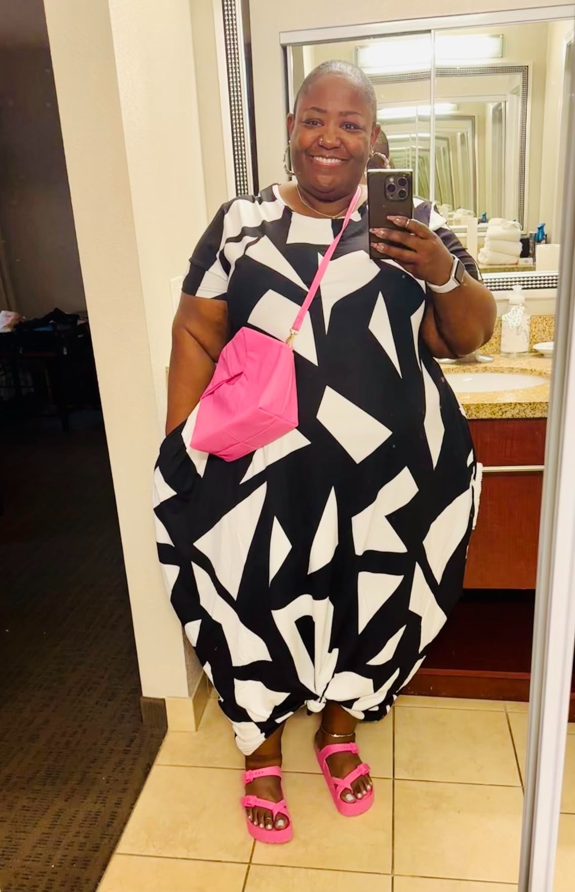 Person successful  a achromatic  and achromatic  geometric formal  with a pinkish  crossbody container  and sandals, taking a reflector  selfie successful  a bathroom