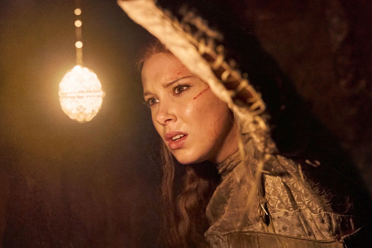 Millie Bobby Brown in "Damsel" in medieval-style clothing looks worried next to a glowing light source in a dim setting