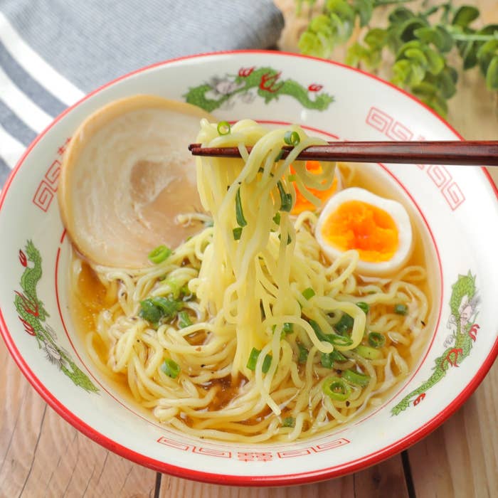 ラーメンの丼に盛られた麺が箸で持ち上げられ、ネギ、半熟卵、チャーシューが添えられている。