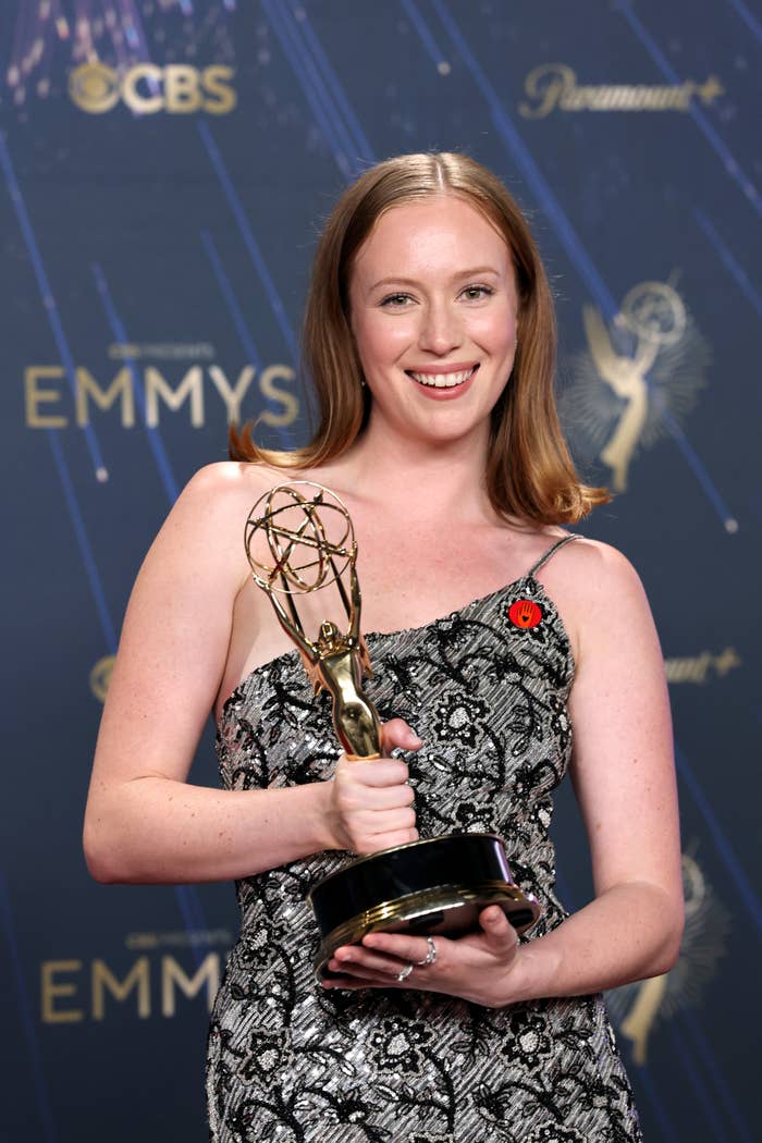 Hannah Einbinder holding an Emmy award, smiling connected the reddish carpet, wearing a one-shoulder patterned formal with a reddish pin