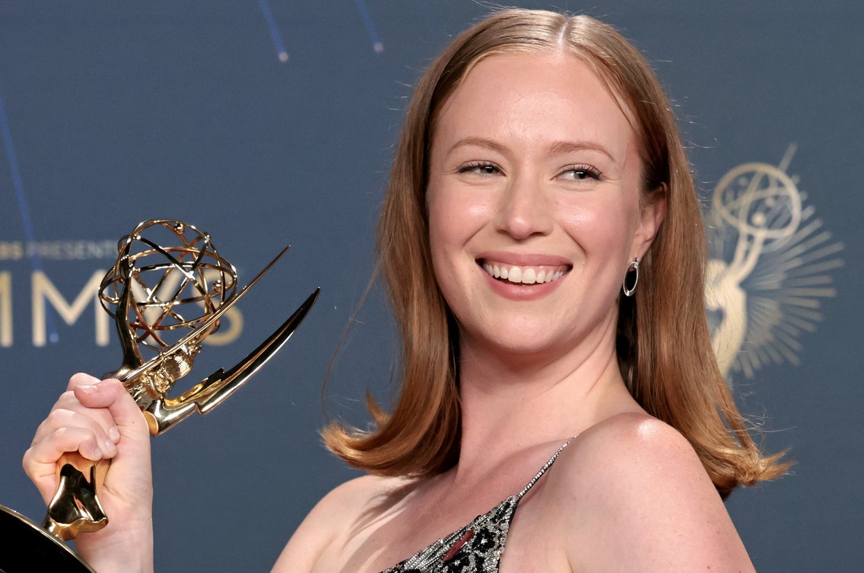 Person in a patterned, sleeveless dress smiling and holding an Emmy award on a blue carpeted stage