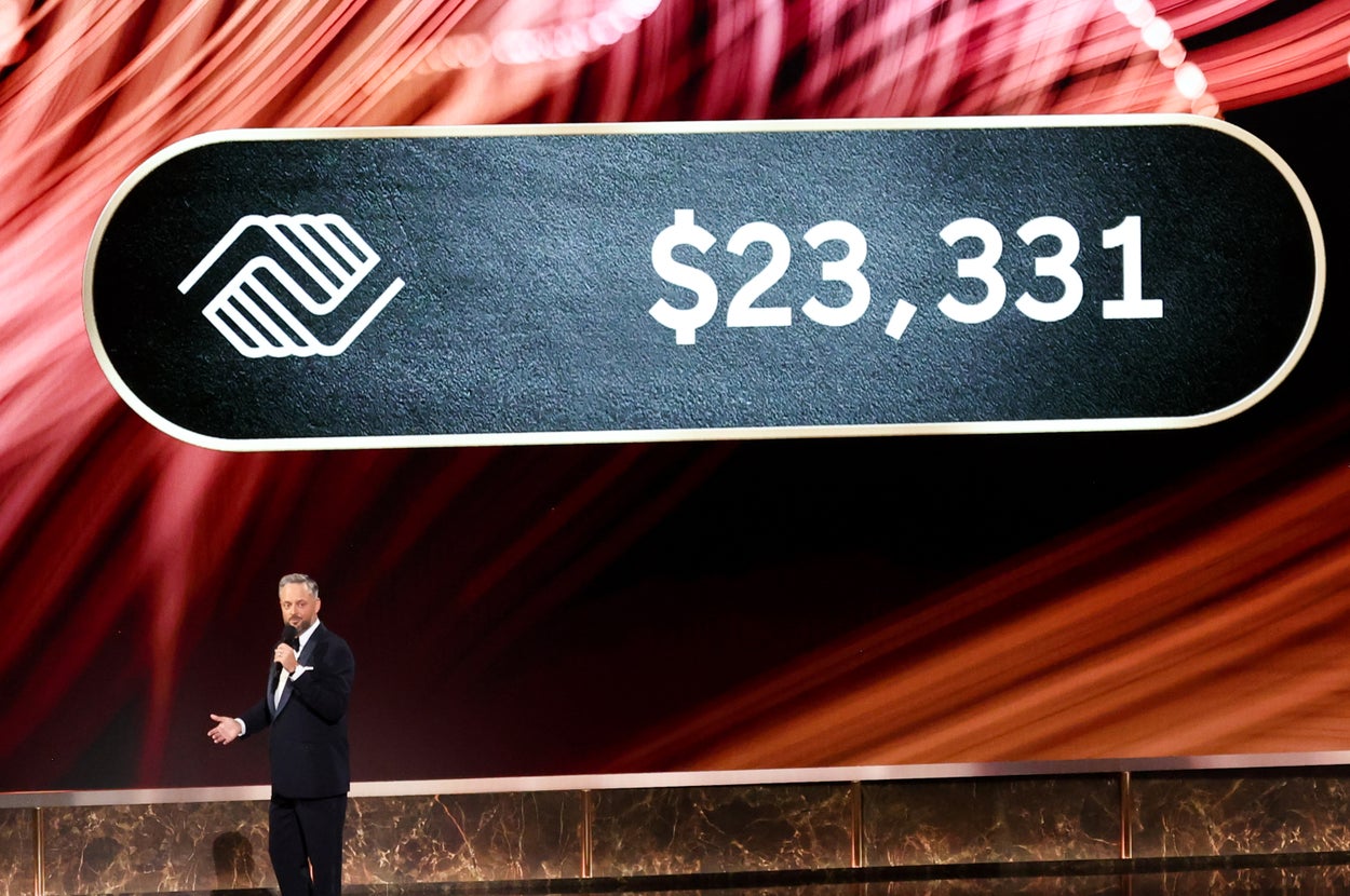 Man in a suit speaks on stage under a sign displaying "$23,331" with an abstract background