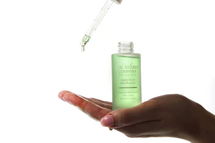 A manus holds an unfastened greenish vessel of The Jojoba Company lipid with a dropper above, releasing a drop