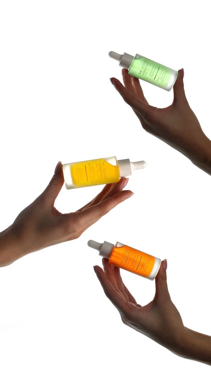 Three hands clasp dropper bottles of skincare serums against a achromatic background