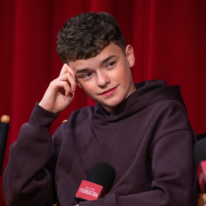 Young person in casual hoodie, sitting on stage with microphone titled "SAG-AFTRA Foundation," looks thoughtful. Red curtain backdrop