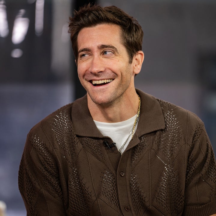 A person smiling in an interview setting, wearing a casual knit shirt over a white tee and a gold chain