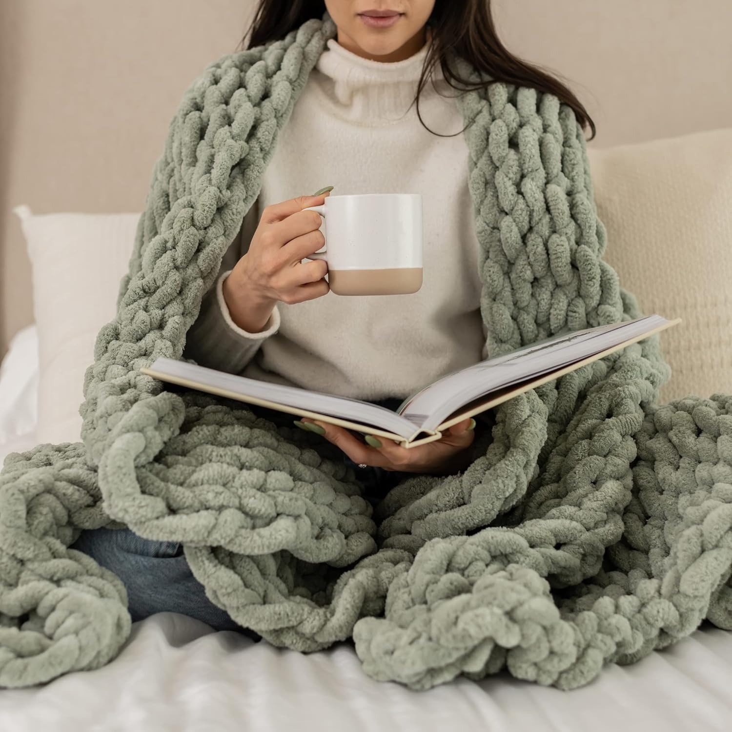 A person sits on a bed, wrapped in a chunky knit blanket, holding a mug, and reading a book, conveying a cozy and relaxed atmosphere