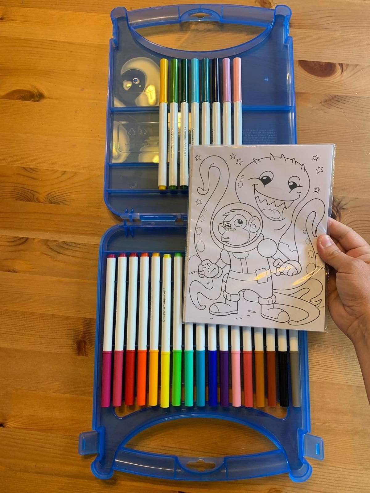 Open art kit with markers and colored pencils, showing a hand holding a coloring page of a child in a spacesuit with a friendly monster