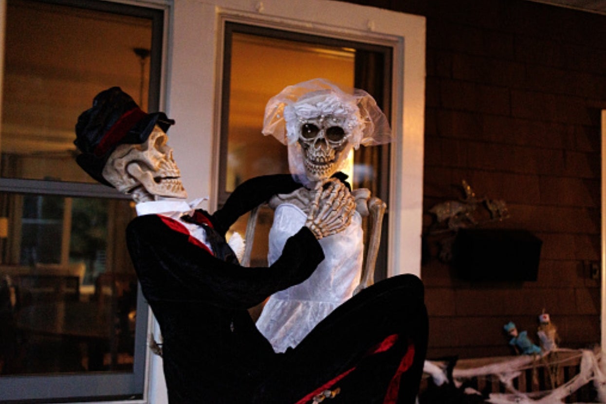Two skeleton figures, dressed as a groom and bride, pose in a humorous wedding scene on a porch