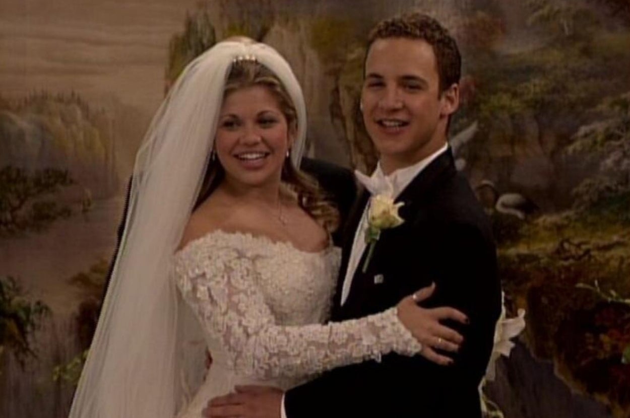 A bride in a lace gown and veil embraces a groom in a tuxedo, both smiling, posing for a formal photo against a painted landscape backdrop