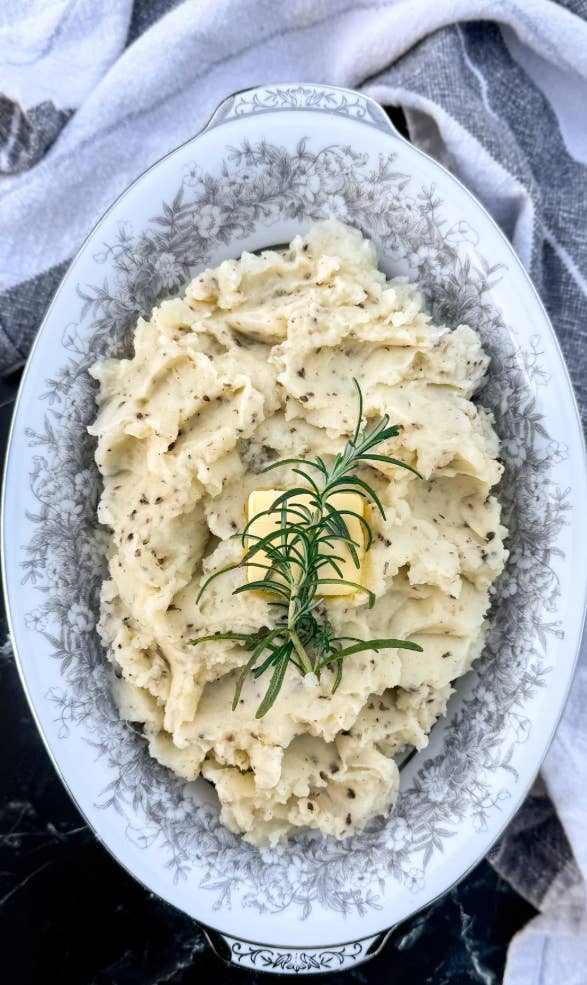 A dish of creamy mashed potatoes garnished with a sprig of rosemary, displayed on a white oval platter