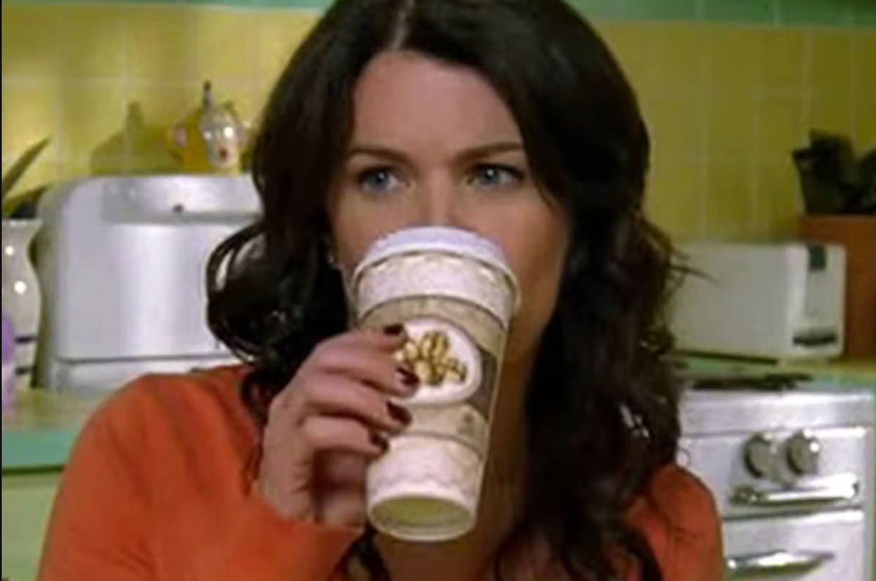Lorelei Gilmore in a kitchen setting drinks from a large coffee cup, wearing a casual sweatshirt