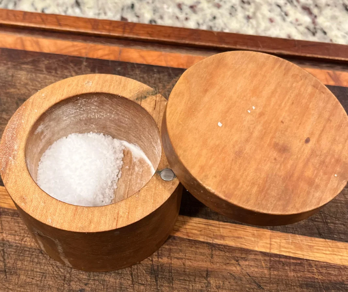 A round wooden salt cellar with a lid partially open, revealing coarse salt inside, displayed on a wooden cutting board