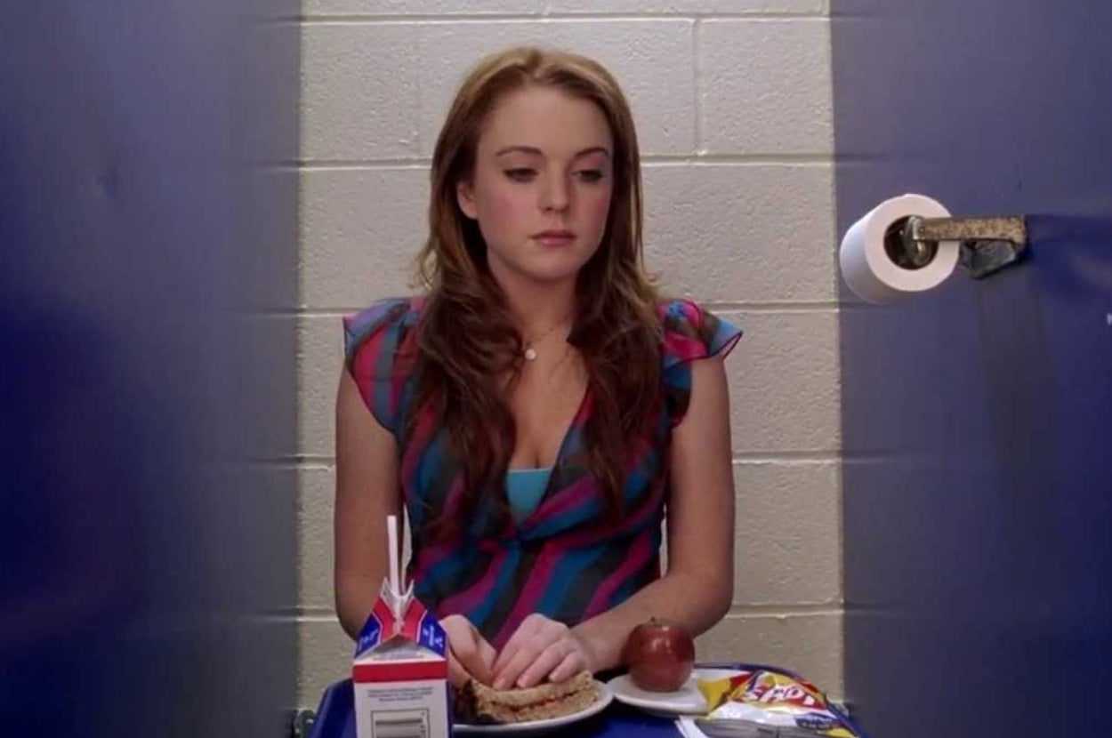 A person sits alone in a bathroom stall with a tray containing a milk carton, sandwich, apple, and chips