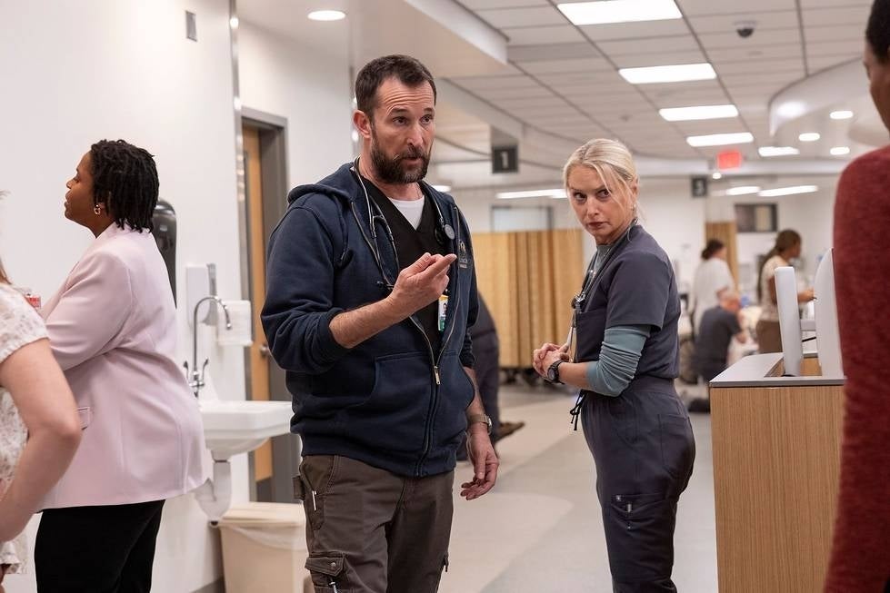 Actors in a hospital setting, one in a hoodie gesturing while another in scrubs listens intently