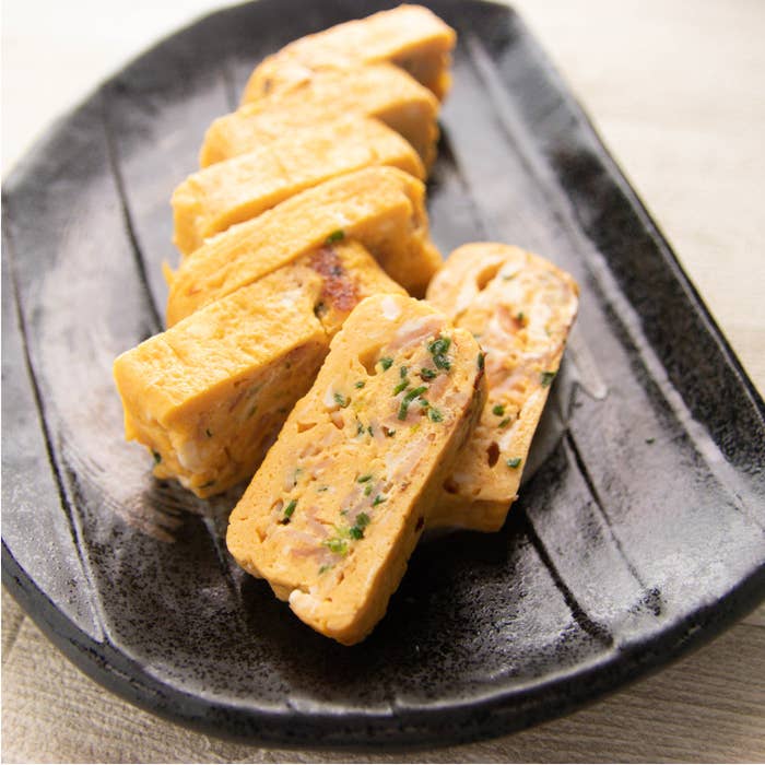 ベーコン入りだし巻き卵。黒い皿に盛られた厚焼き卵ロール。薬味が混ざり、食欲をそそる仕上がり。