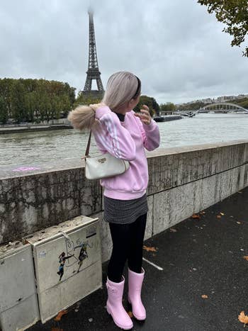 Person successful a stylish casual outfit with pinkish boots looks astatine the Eiffel Tower, lasting adjacent a river