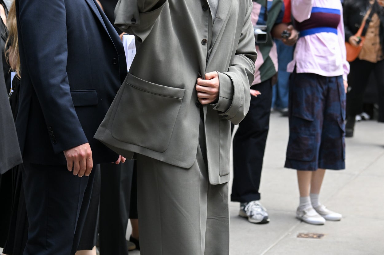 Person in an oversized suit waves while walking outside, surrounded by people and security. Fans and photographers capture the moment