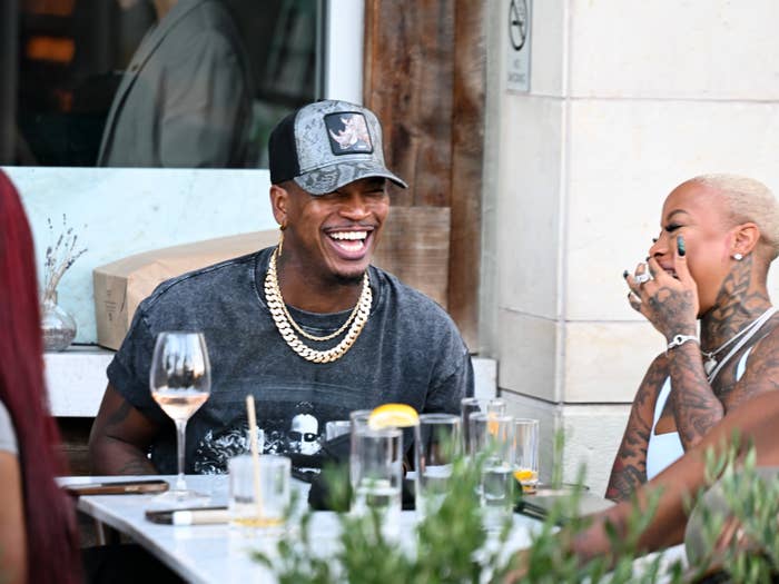 Two radical laughing portion eating outdoors; 1 wears a headdress and concatenation necklaces, the different has tattoos and shaved hair. Casual and joyful atmosphere