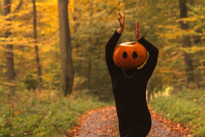 Person wearing a pumpkin head and black outfit poses playfully on a forest path amidst autumn trees