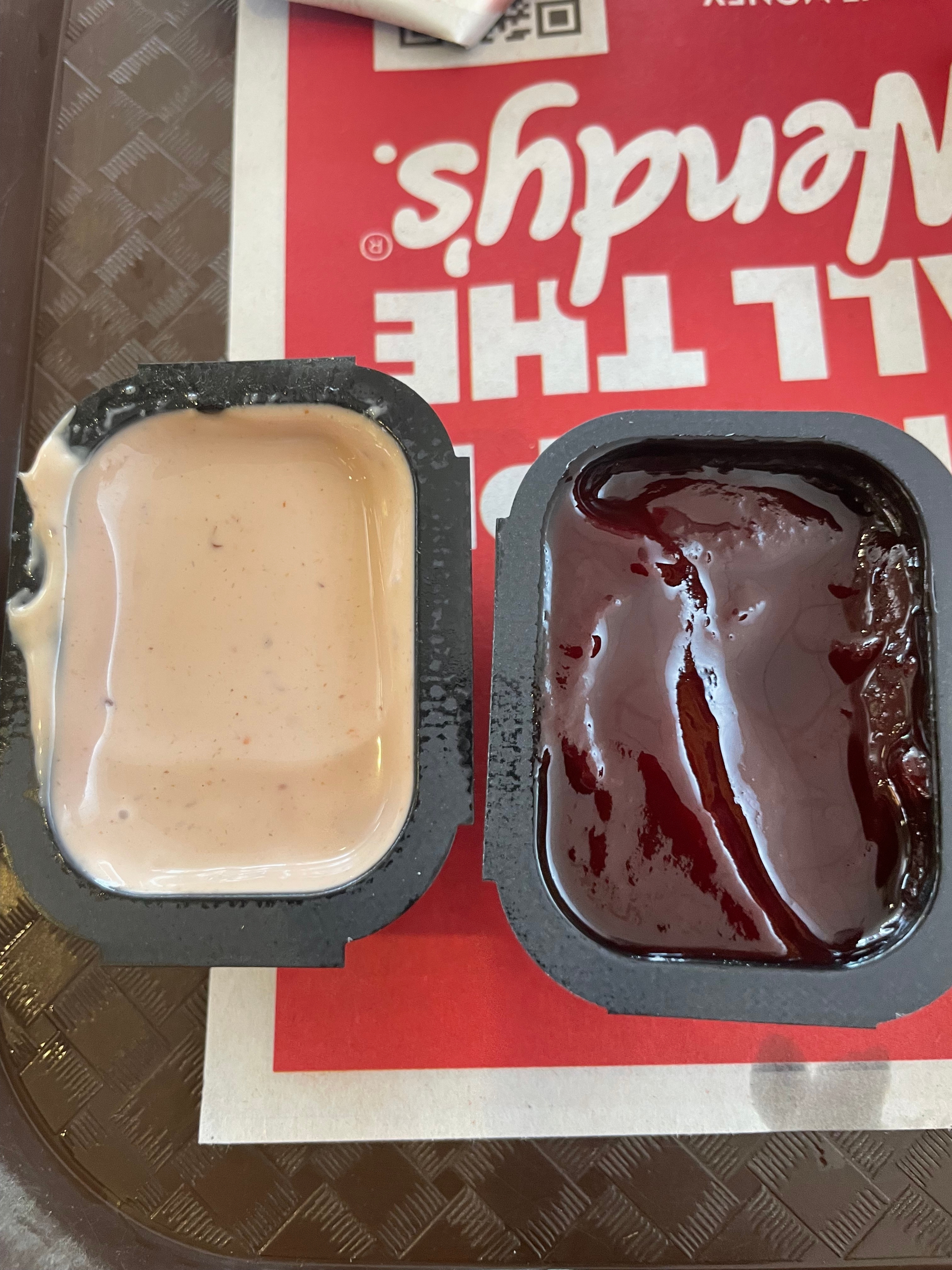 Two dipping sauces, one creamy, the other glossy and dark, in small containers on a tray. A Wendy's logo is partially visible in the background