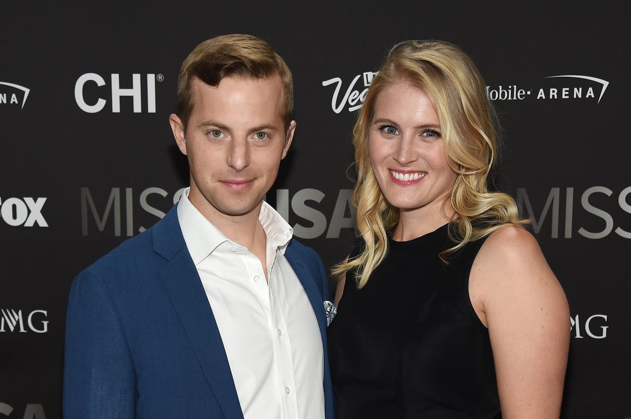 A couple poses at a media event. The man wears a suit, and the woman wears a floral skirt and sleeveless top