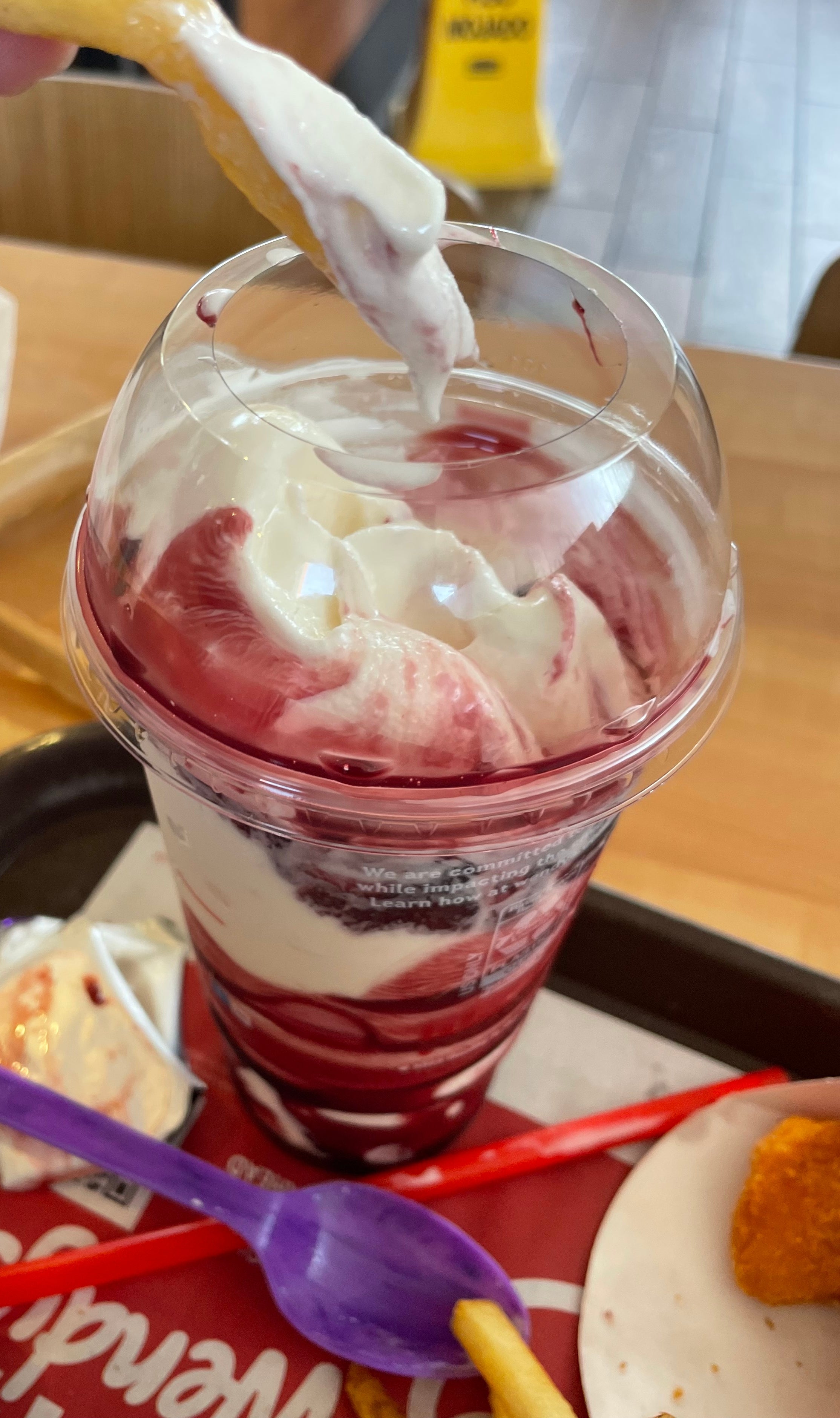 A person dips a French fry into a berry sundae inside a fast food restaurant. A tray with a wrapper and utensils is visible