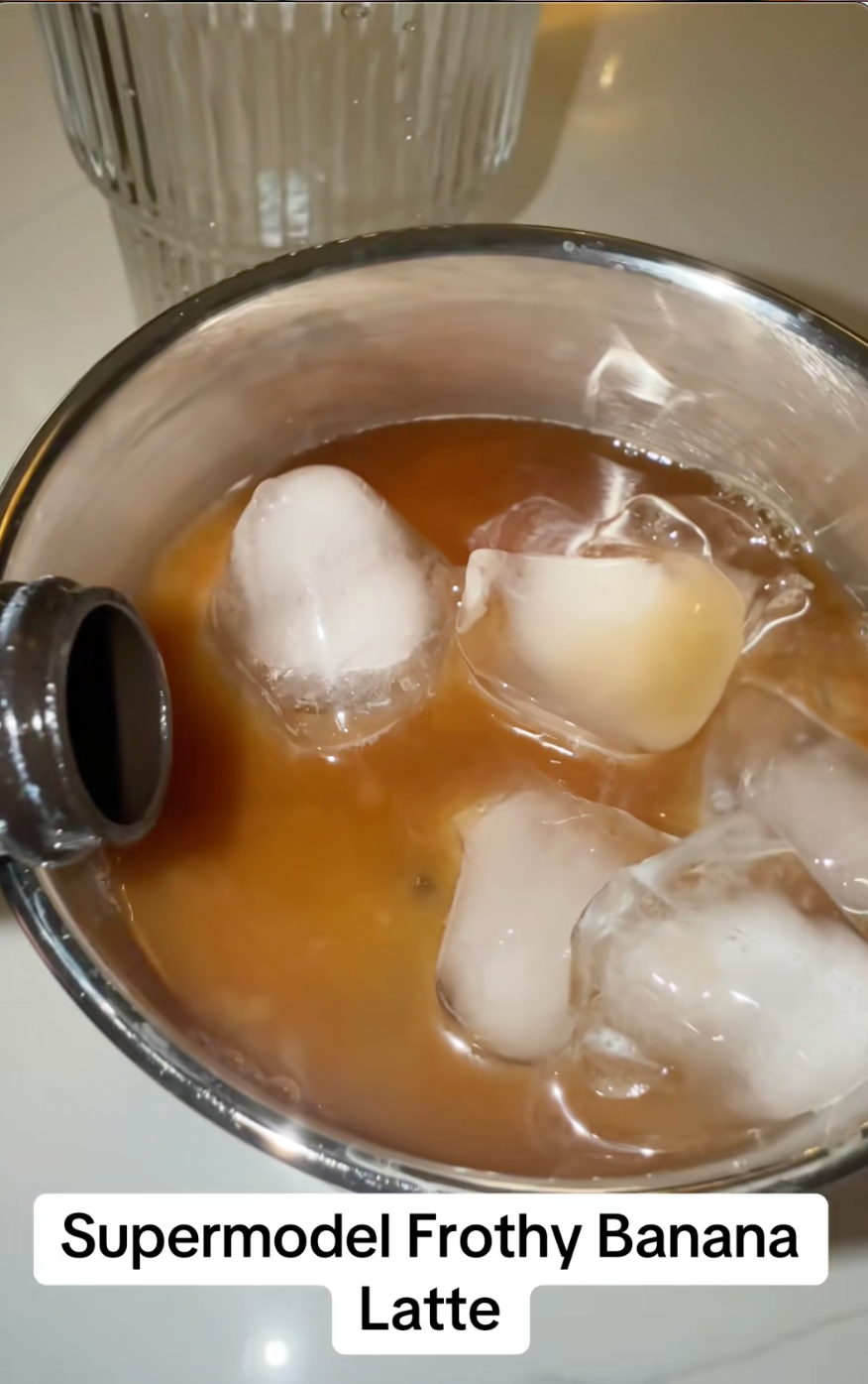 Iced banana latte in a tumbler with large ice cubes, bottle spout nearby. Text: "Supermodel Frothy Banana Latte."