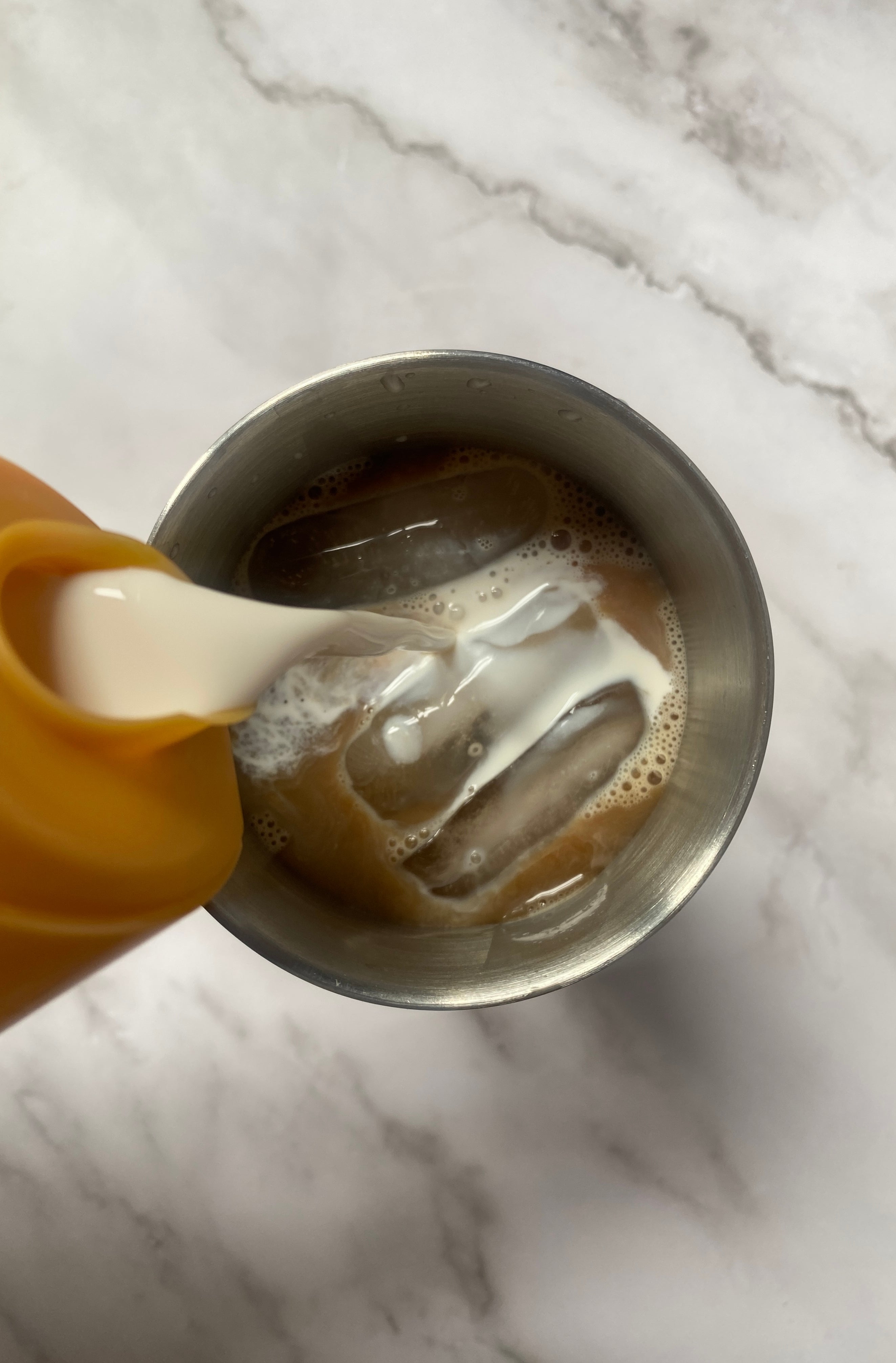 Coffee creamer is being poured from a yellow container into a metal cup with iced coffee