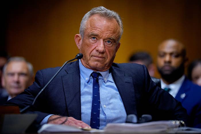 A person in a suit sits at a desk with a microphone in a formal setting, looking serious during what appears to be a hearing or conference