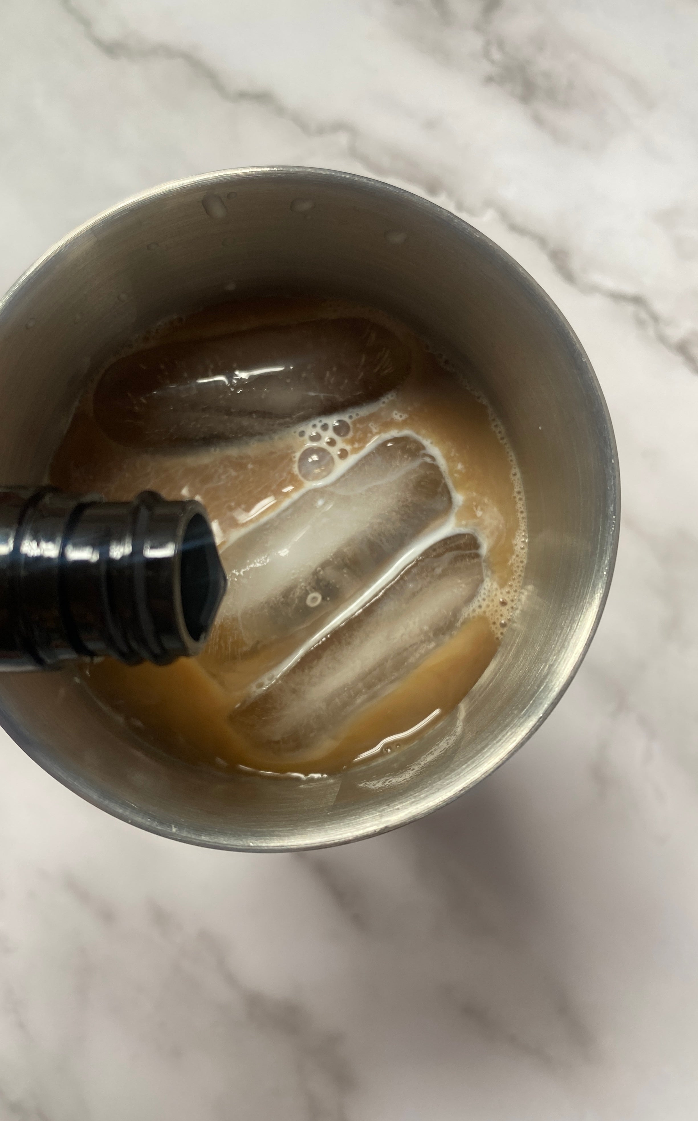 Iced coffee in a stainless steel cocktail shaker with a hand holding a bottle pouring liquid into it
