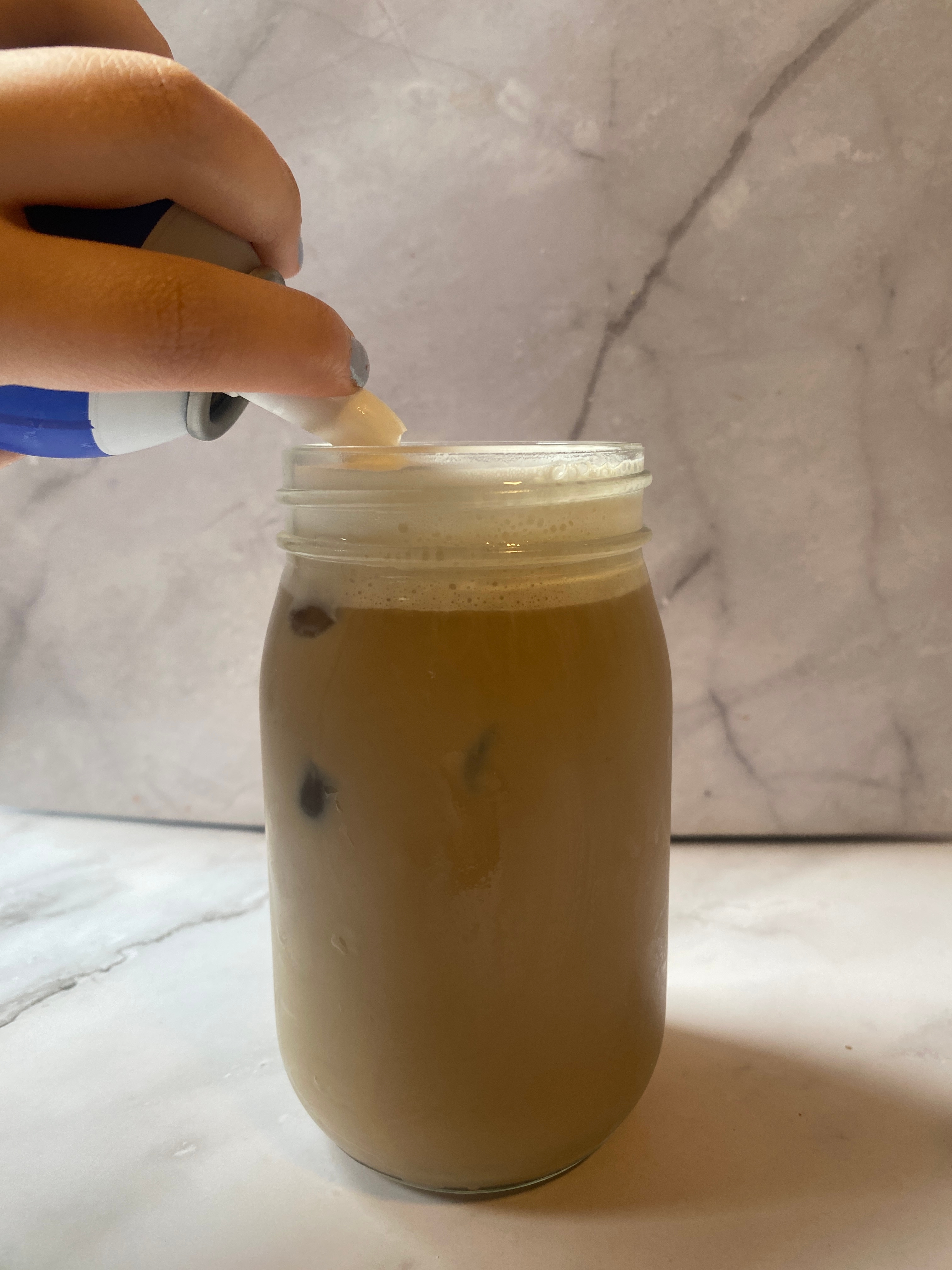 A hand is adding whipped cream to an iced coffee in a glass jar on a marble surface