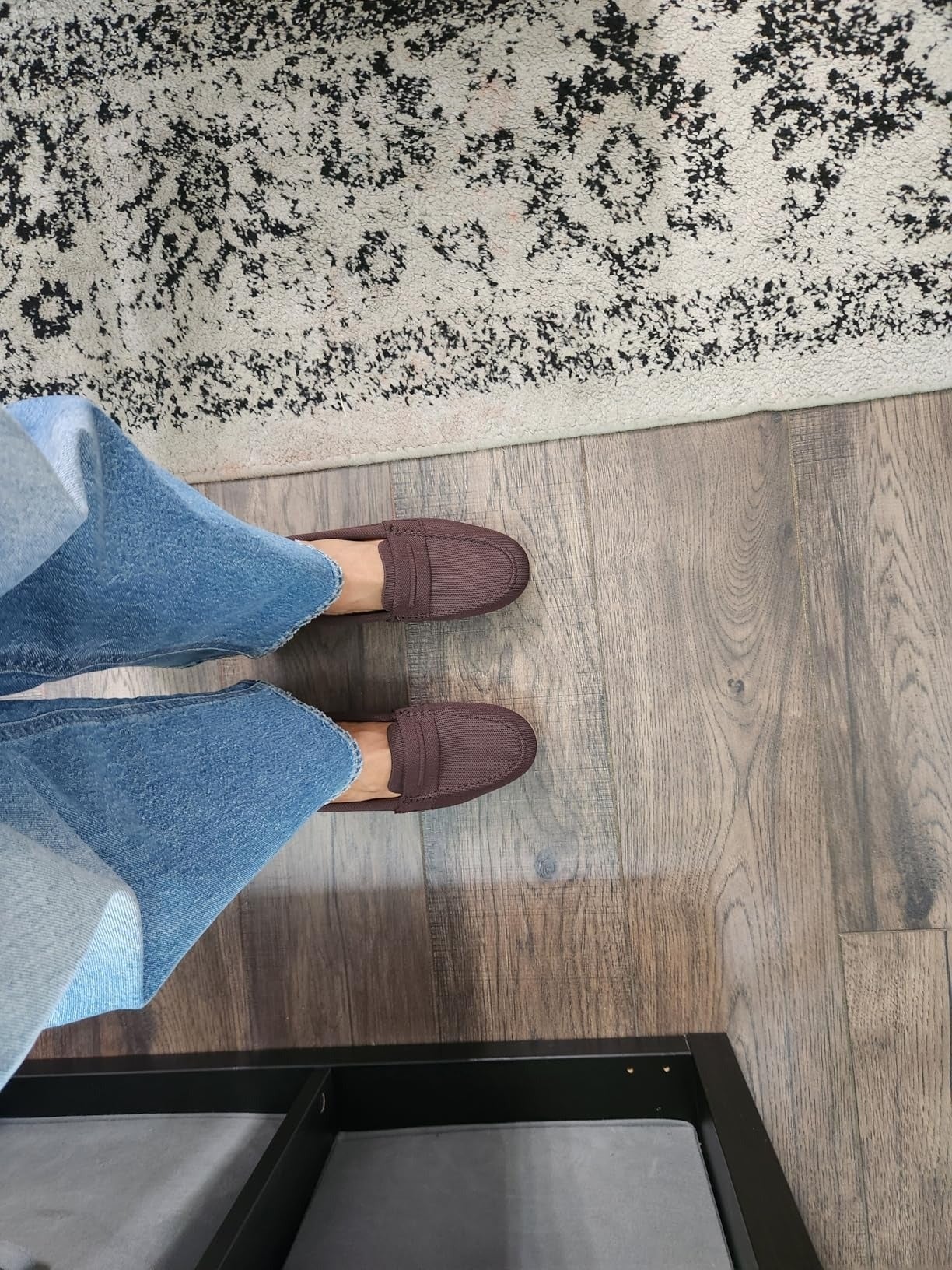 Person wearing jeans and brownish  loafers stands connected  a woody  level  adjacent  to a patterned rug