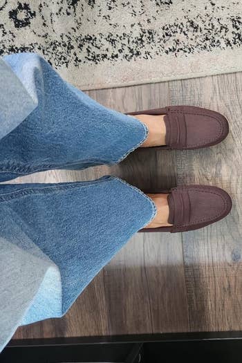 Person wearing jeans and brownish loafers stands connected a woody level adjacent to a patterned rug