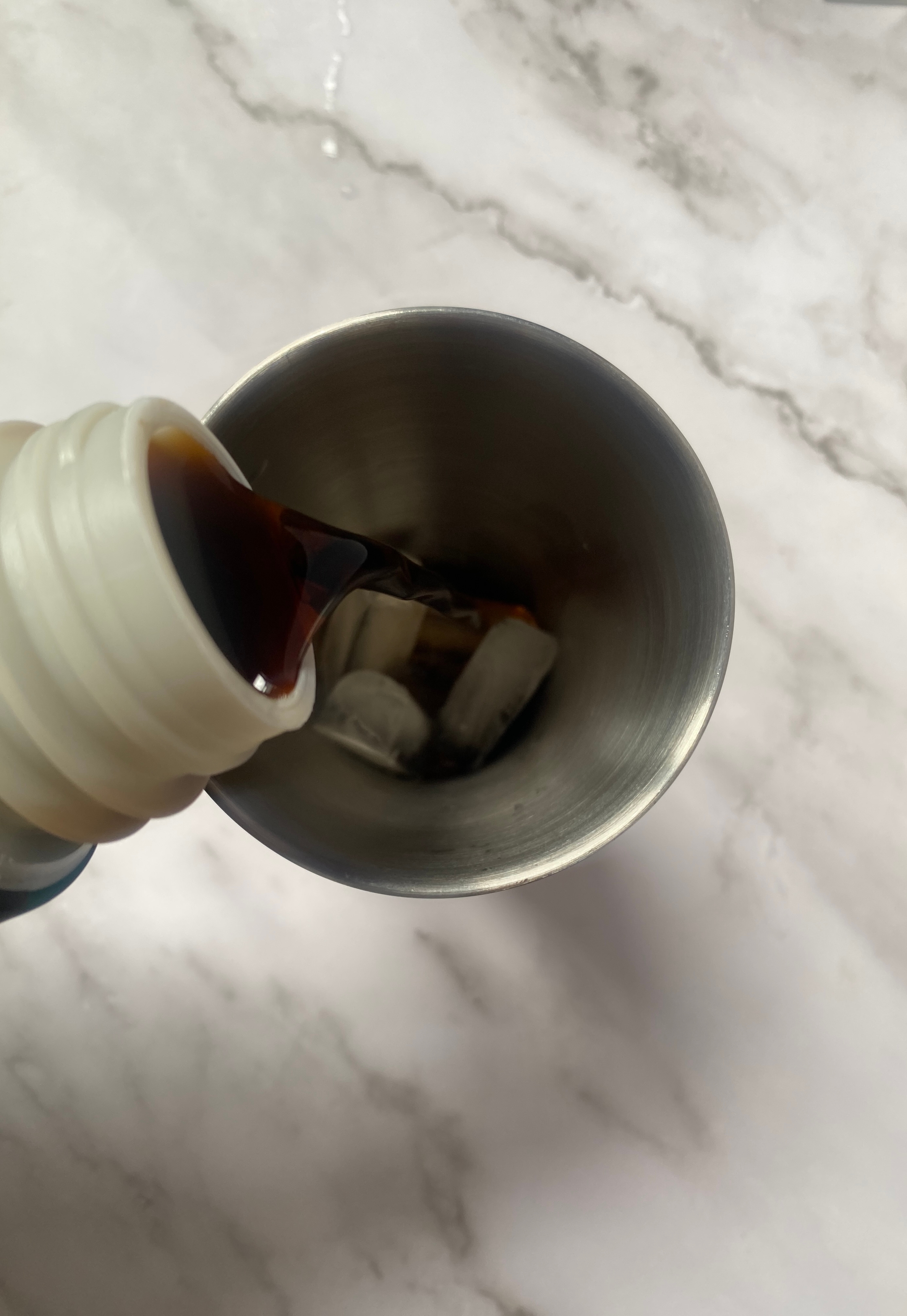 Pouring dark liquid into a metal cup with ice on a marble surface